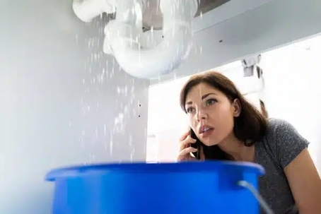 A woman with a concerned expression, calls on a smartphone, as water leaks from exposed pipes into a blue bucket.