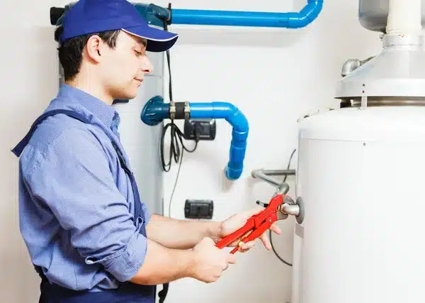A plumber wearing a blue uniform is repairing a water heater, using a red wrench on a valve.